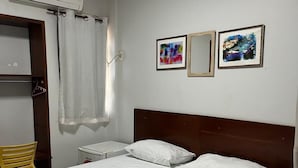 A neatly made bed with white linens, a wooden headboard, and a plastic bag on the bed. There is a small bedside table with a lamp, a chair, and a window with curtains. Two framed pictures hang on the wall above the bed. - Mais Hotel (Araguaina)