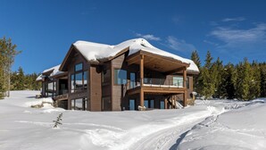 A modern log cabin with multiple large windows and a balcony, surrounded by snow-covered landscape and trees. - Silver Aspen | Fire Pit, Hot Tub & Views (Big Sky)