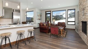 A modern kitchen with a central island, bar stools, and a seating area with a fireplace. - Silver Aspen | Fire Pit, Hot Tub & Views (Big Sky)