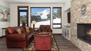 A living room with a fireplace, leather sofa, armchair, and large windows with a snowy mountain view. - Silver Aspen | Fire Pit, Hot Tub & Views (Big Sky)