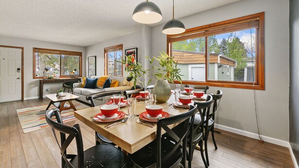 A dining area with a wooden table set for four, surrounded by black chairs. There are two pendant lights above the table, and a large window with a view of neighboring houses. - Old Town Basecamp 6 Blocks to Old Town Square (Fort Collins)