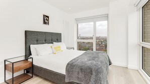 A modern bedroom with a large bed, a glass side table, a view of the city through a large window, and a cityscape visible outside.
