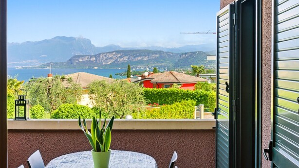 A balcony with a table, chairs, a potted plant, and a view of a lake and mountains. - Regarda -Blue View 3  Apartment with lake view and pool (Bardolino)