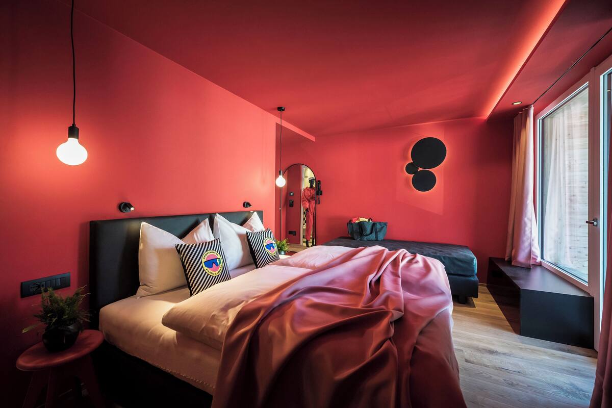 Une chambre moderne avec un mur rouge, un lit agrémenté de plusieurs coussins, une table de chevet et une fenêtre avec des rideaux.