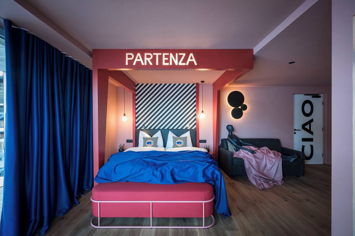 Une chambre moderne avec un lit, une tête de lit à motifs rayés, un mur d’accent rouge et un luminaire fixé au mur.