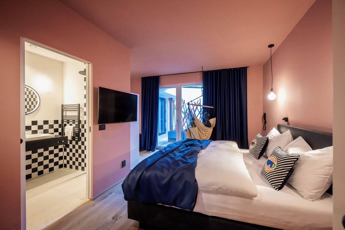 Une chambre d’hôtel avec une salle de bain aux carreaux, un lit avec une literie à rayures et un téléviseur fixé au mur.