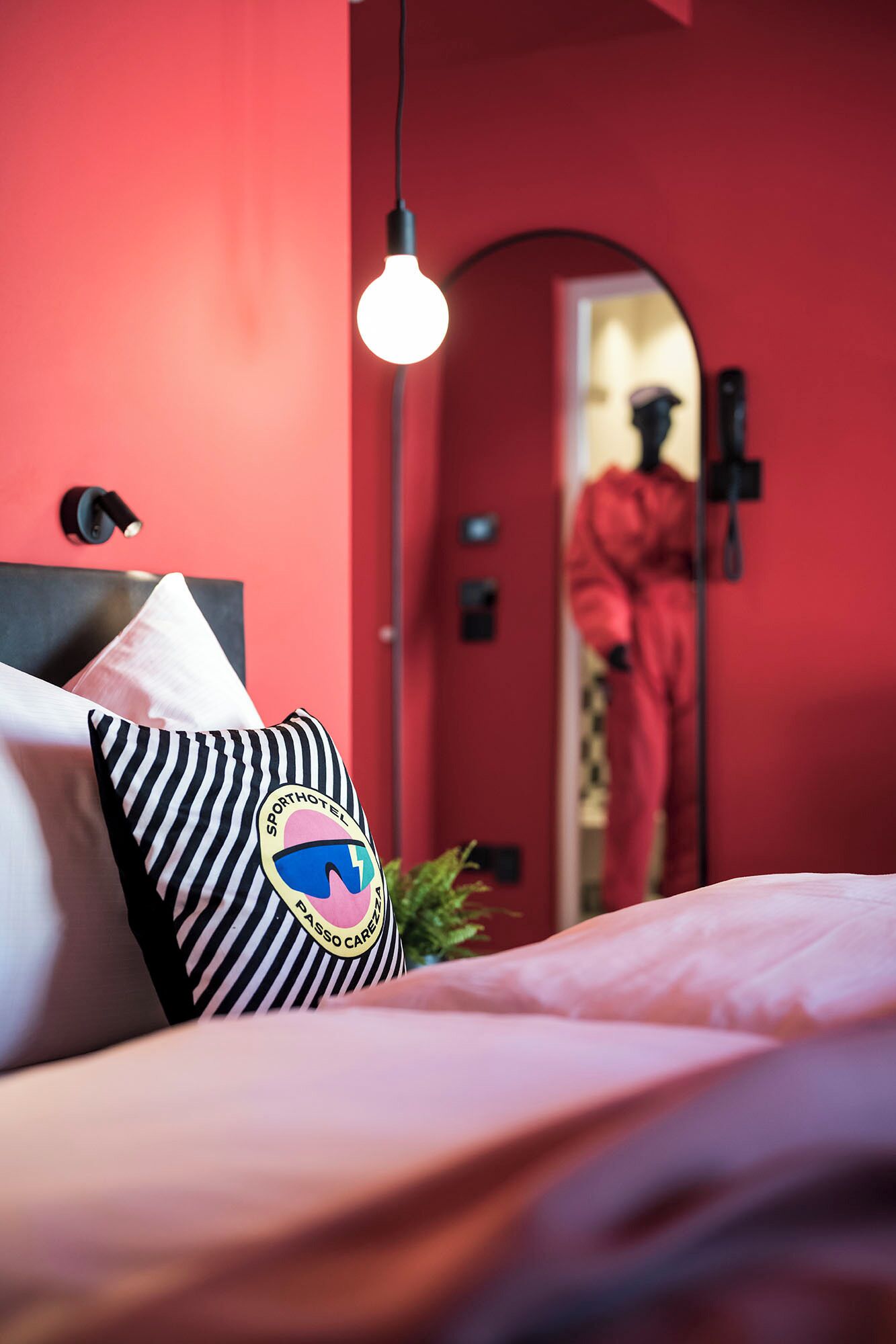 Ein Bett mit einem gestreiften Kopfkissen und einem Logo, eine hängende Glühbirne und eine rote Wand.