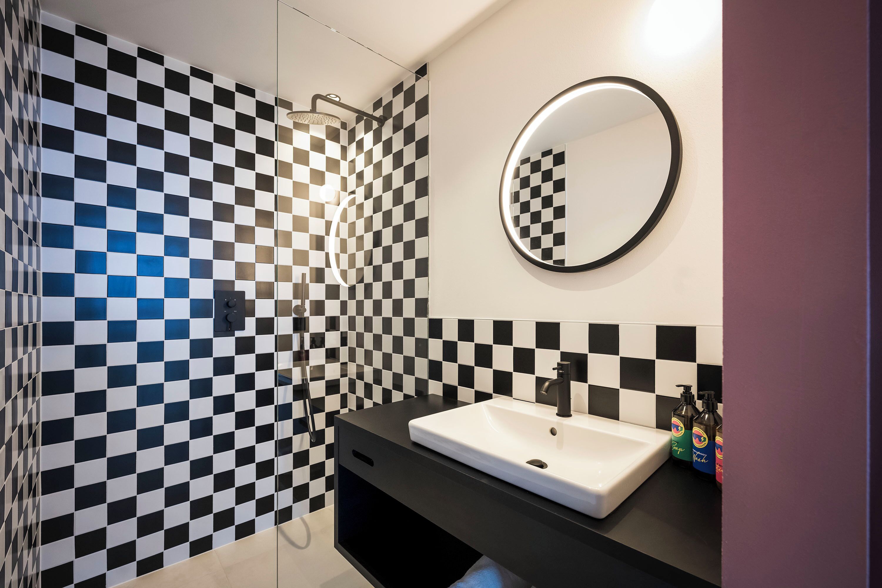 Ein Badezimmer mit Wand aus schwarz-weißen Schachfeldern, einem weißen Waschbecken, einem runden Spiegel und einer Duschkabine.