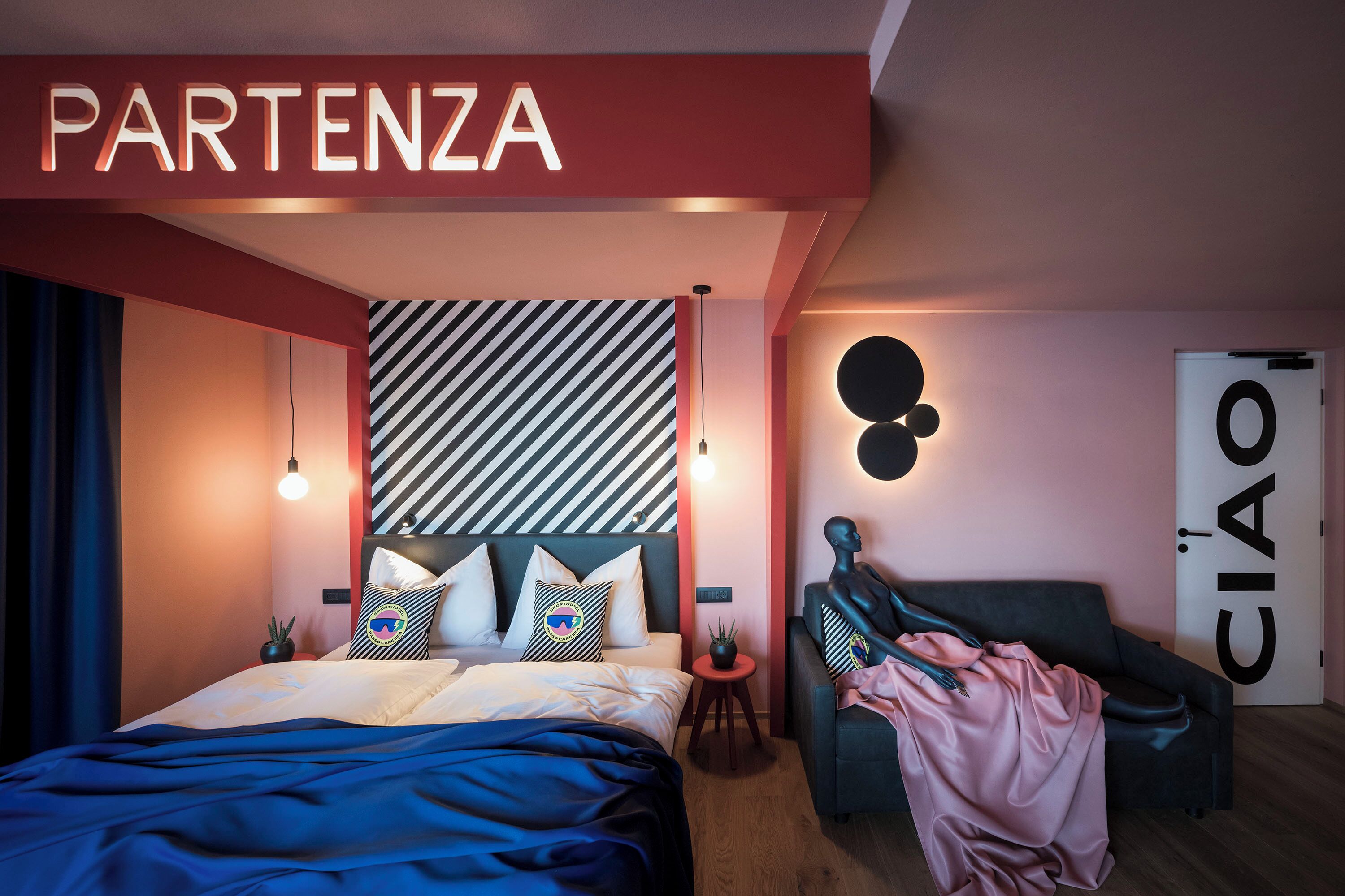 A modern bedroom with a bed, a sofa, a wall-mounted light, and a 'PARTENZA' sign.
