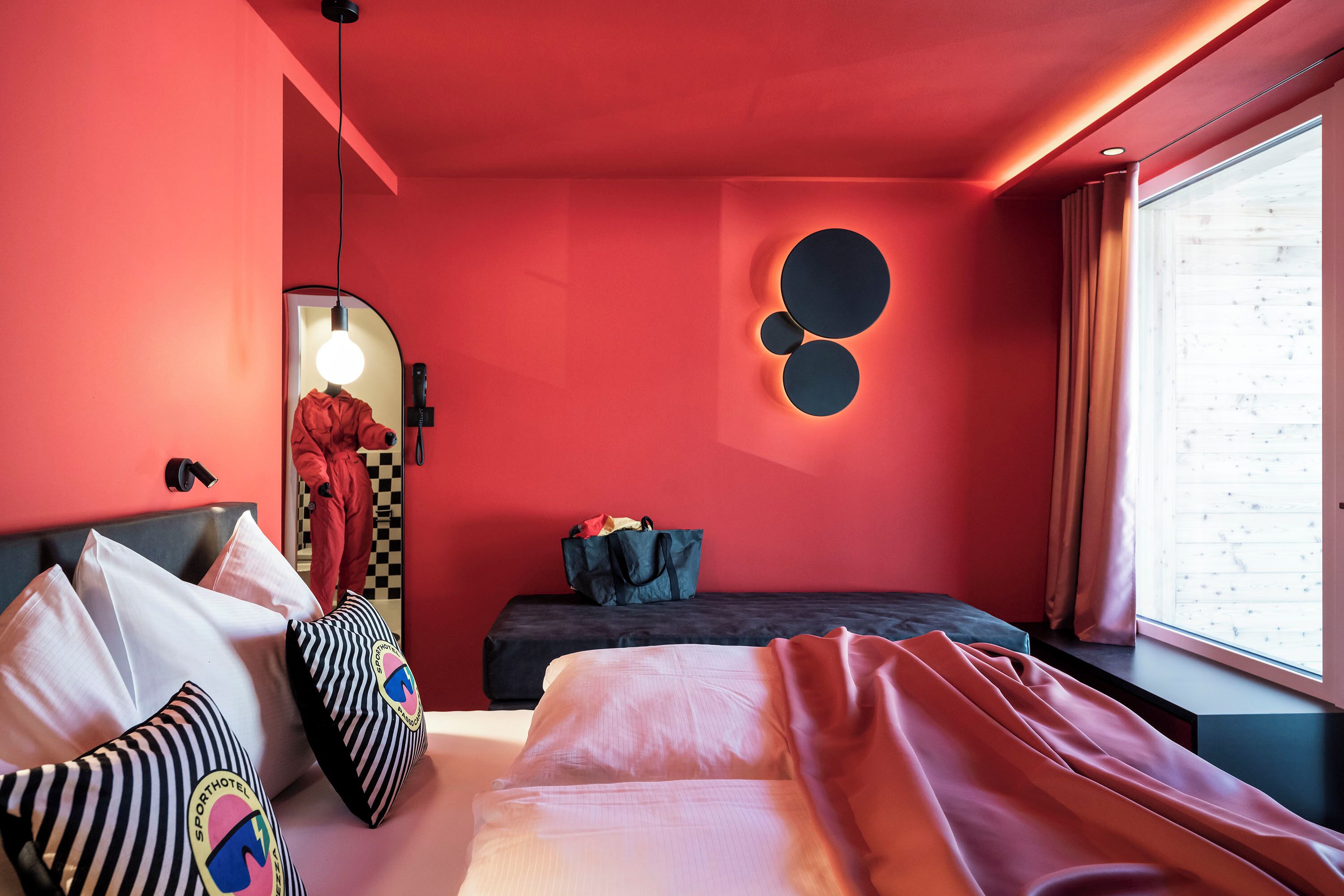Habitación de hotel moderna con una pared roja, cama con ropa de cama blanca, sofá negro y una ventana grande con cortinas.