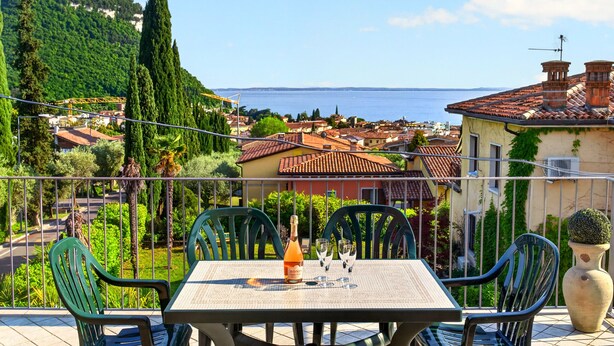 A balcony with a table set for two, overlooking a coastal town with houses and greenery. - Regarda - La Terrazza di Garda lake view apartment with terrace (Garda)