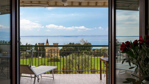 A balcony with a view of a castle, a green lawn, and a body of water.