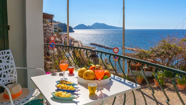 Ein Balkon mit Esstisch, der Blick auf die Küste und in die Ferne zu den Bergen gewährt.