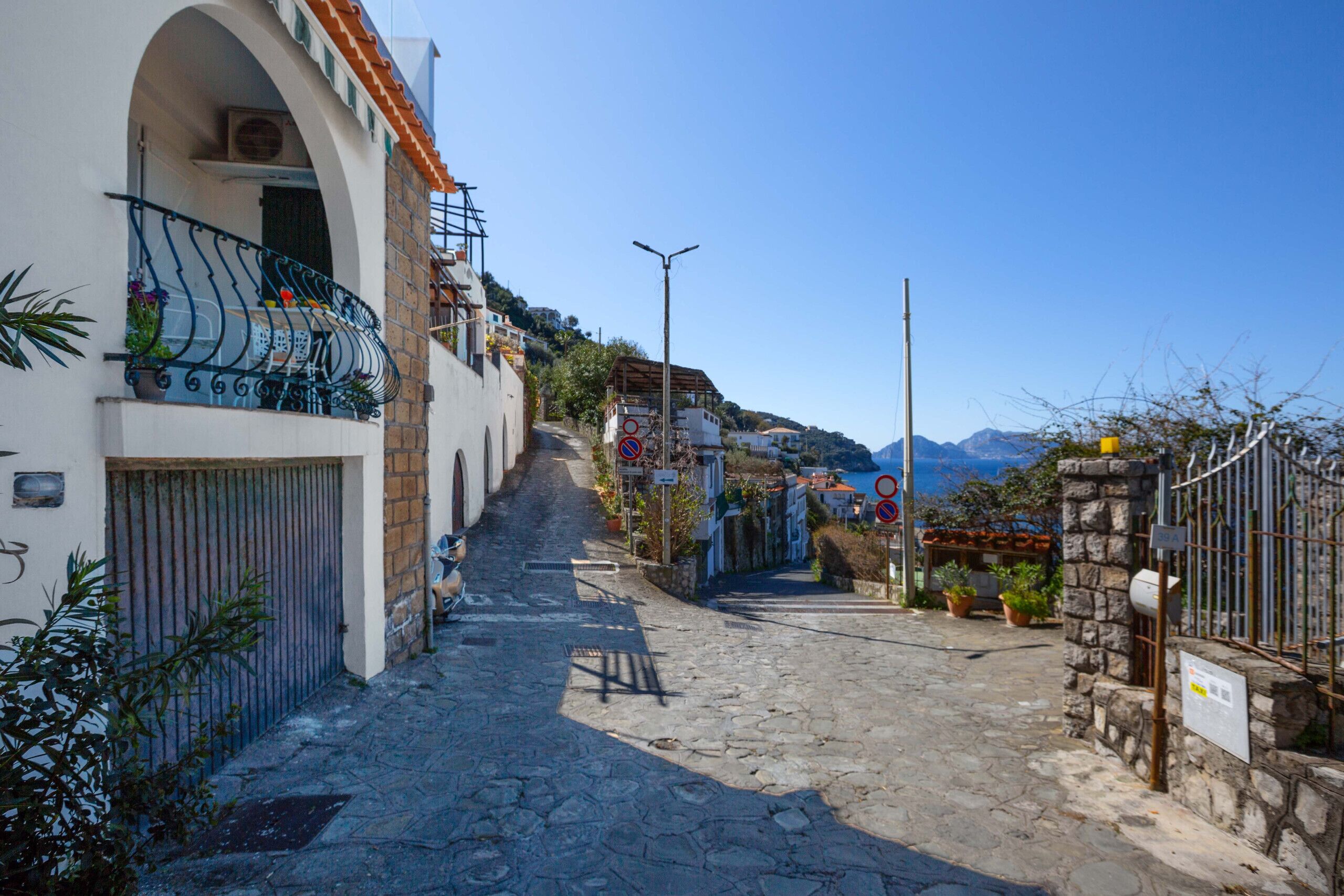 Une ruelle étroite pavée, bordée d’immeubles dotés de balcons, un ciel dégagé et des montagnes en arrière-plan.