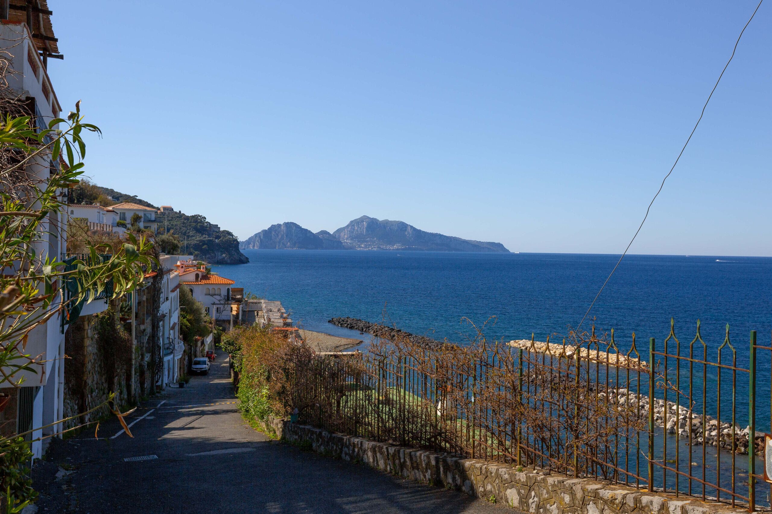 Une rue étroite qui mène à une zone côtière, d’où l’on peut admirer la mer et les montagnes en arrière-plan.