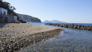 Ein Kiesstrand mit klarem Wasser, eine felsige Küste und ein Gebäude am Hang.