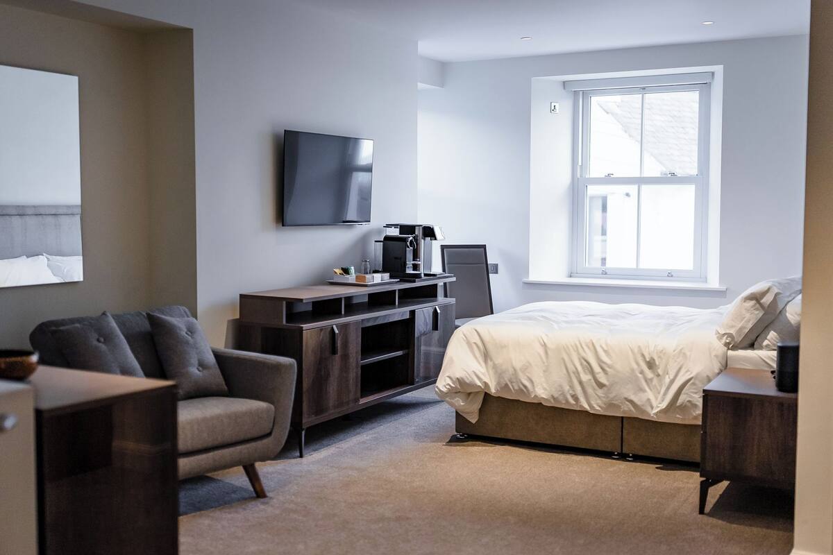 Ein modernes Hotelzimmer mit einem Bett, einem Schreibtisch mit Computer, einem Sessel, einem Fernseher und einem Fenster.