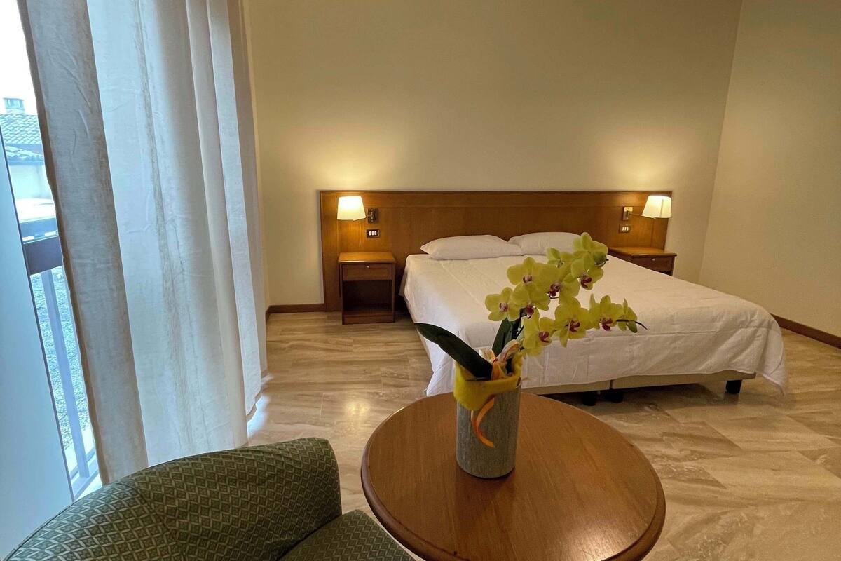 A hotel room with a bed, a nightstand with a lamp, a green patterned armchair, a round wooden table with a vase of flowers, and a hanging light fixture.
