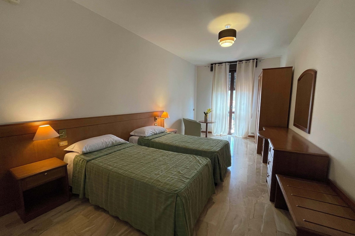 A hotel room with two beds, a desk, a chair, a small table with a lamp, and a large mirror.