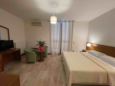 A hotel room with a bed, a TV, a small table with a plant, a chair, and a desk with a computer.