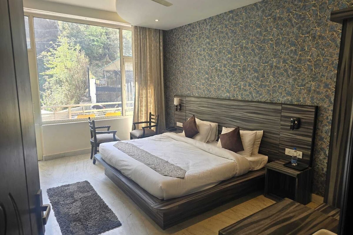 Um quarto com uma cama grande, uma mesa de cabeceira, uma cadeira, uma janela com cortinas e vista para árvores e um edifício lá fora.