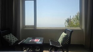 A room with a large window, a bed, a glass-top table, two chairs, and a view of greenery outside.