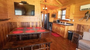 A wooden cabin interior with a dining table, benches, a kitchen area with appliances, and a television mounted on the wall. - Secluded Cabin in Weatherford, TX (Weatherford)