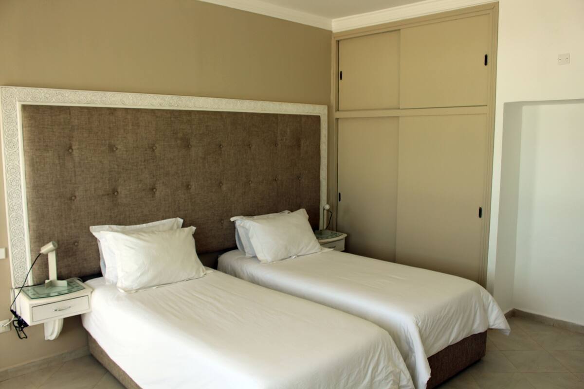 Une chambre d’hôtel avec deux lits simples, une grande tête de lit rembourrée, une table de chevet avec une lampe et une armoire.