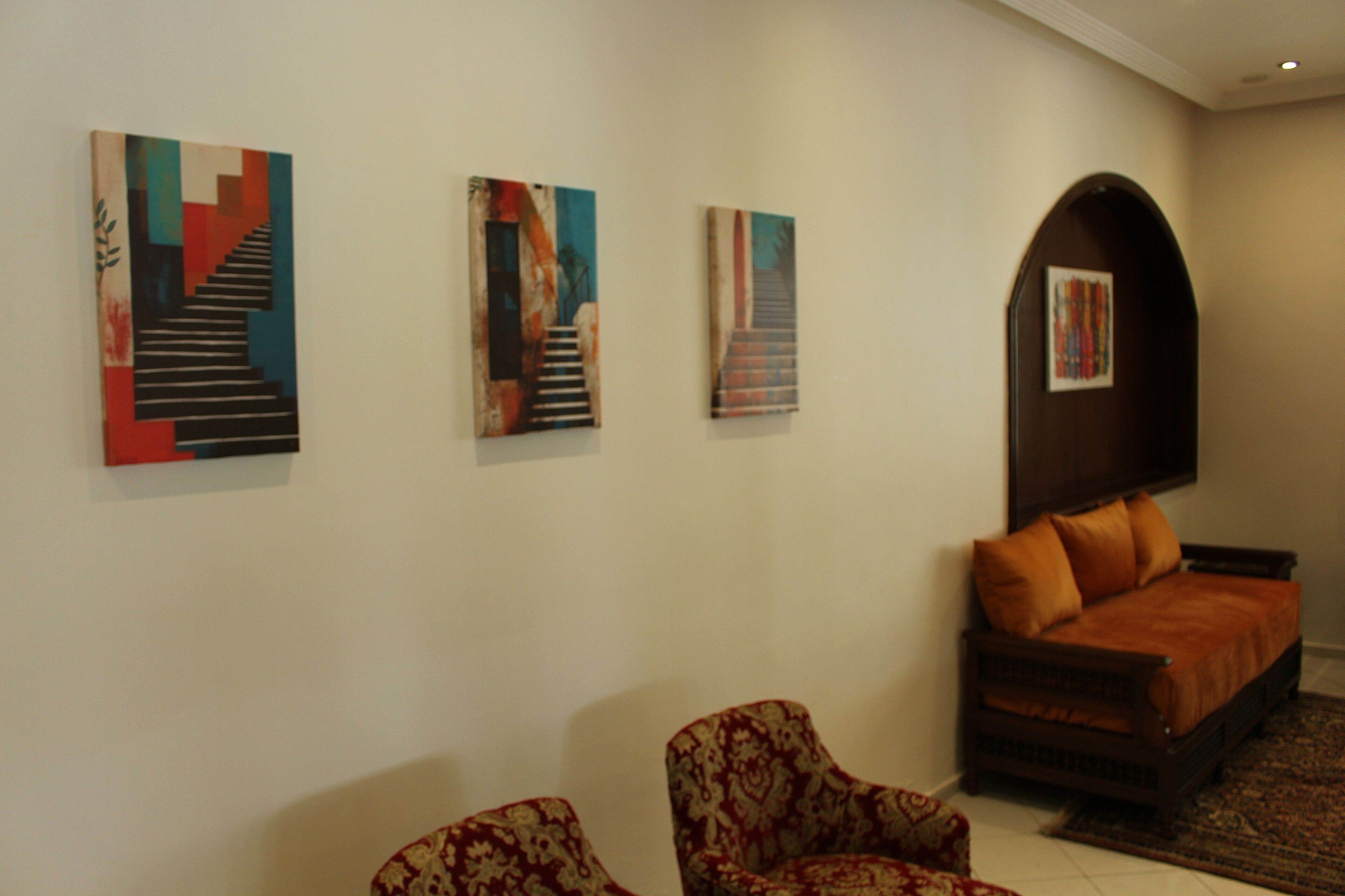 A room with three abstract paintings on the wall, a sofa with orange cushions, and two patterned armchairs.