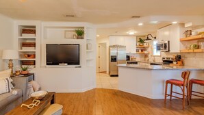 A modern kitchen with white cabinets, a built-in island with bar stools, a large flat-screen TV, and a dining area with a wooden bench.