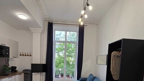A modern room with a bed, a blue sofa, a black wardrobe, a kitchen area, and a large window with curtains. - Studio Centre Paris  Opera (Paris)