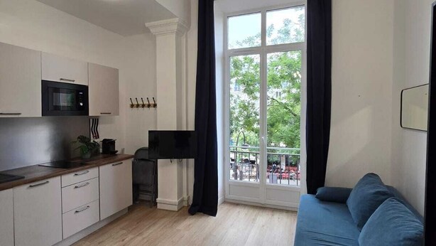 A modern living room with a blue sofa, a kitchen area with a microwave, and a large window with curtains. - Studio Centre Paris  Opera (Paris)