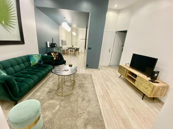 Een moderne woonkamer met een groene bank, een ronde salontafel, een tv op een houten dressoir, en een eethoek op de achtergrond.