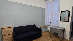 Una habitación con un sofá de color oscuro, una mesita de noche de madera, una mesa gris y una ventana con persianas.