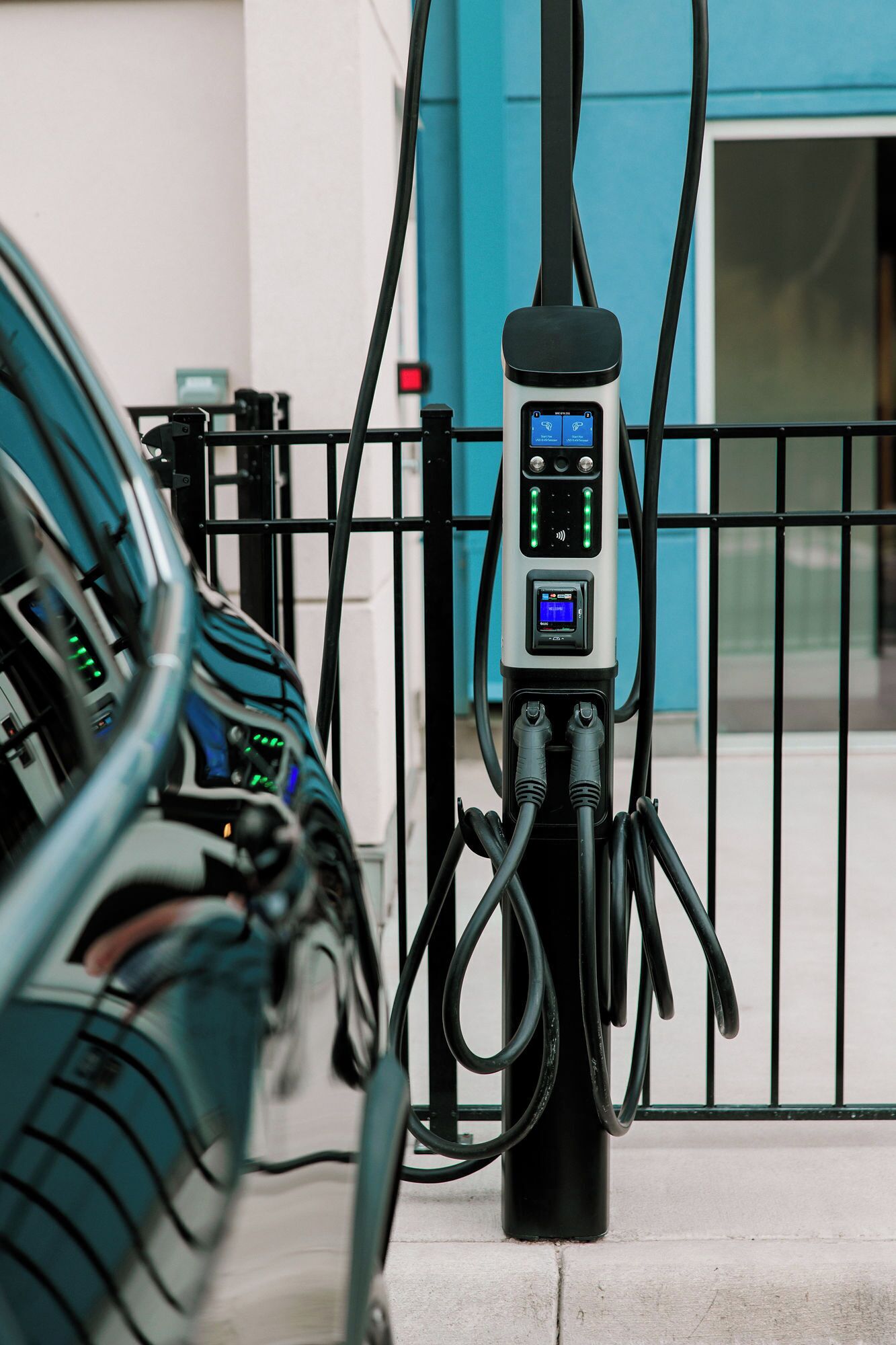 A charging station with multiple cables connected to it, set against a building with a blue door and a black metal fence.