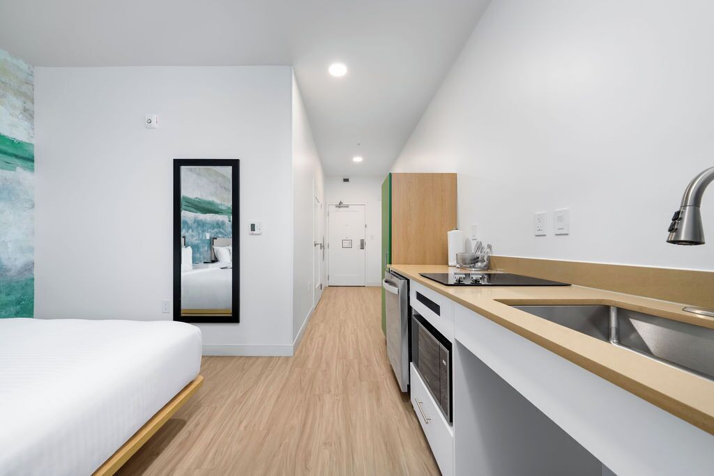 A modern hotel room with a bed, a kitchenette, a sink, and a door leading to another room.