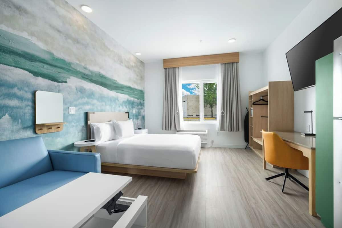 A modern hotel room with a bed, a desk, a chair, a TV, a sofa, and a large window with curtains.