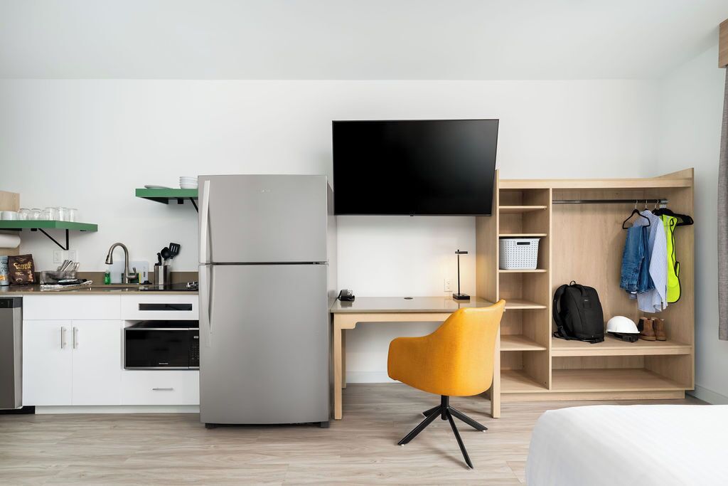 A compact hotel room with a kitchenette, a flat-screen TV, a desk, a chair, and a wardrobe.