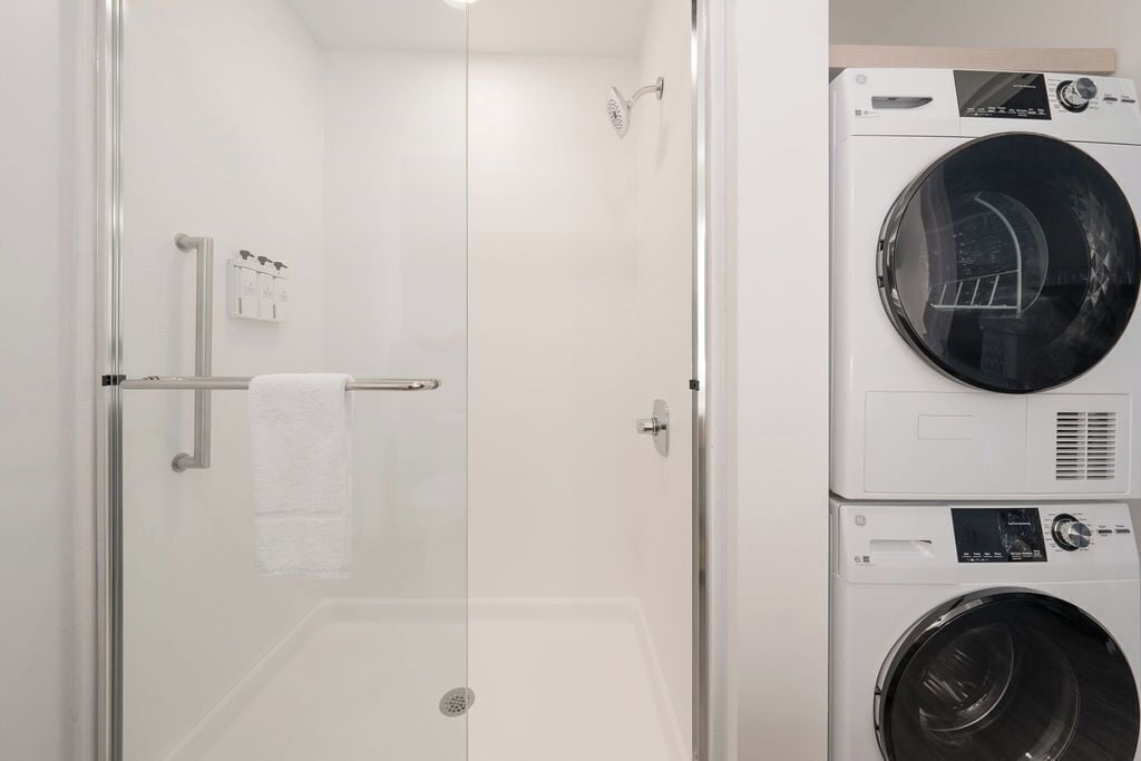 A hotel bathroom with a glass-enclosed shower, a stacked washer and dryer, and a towel rack.