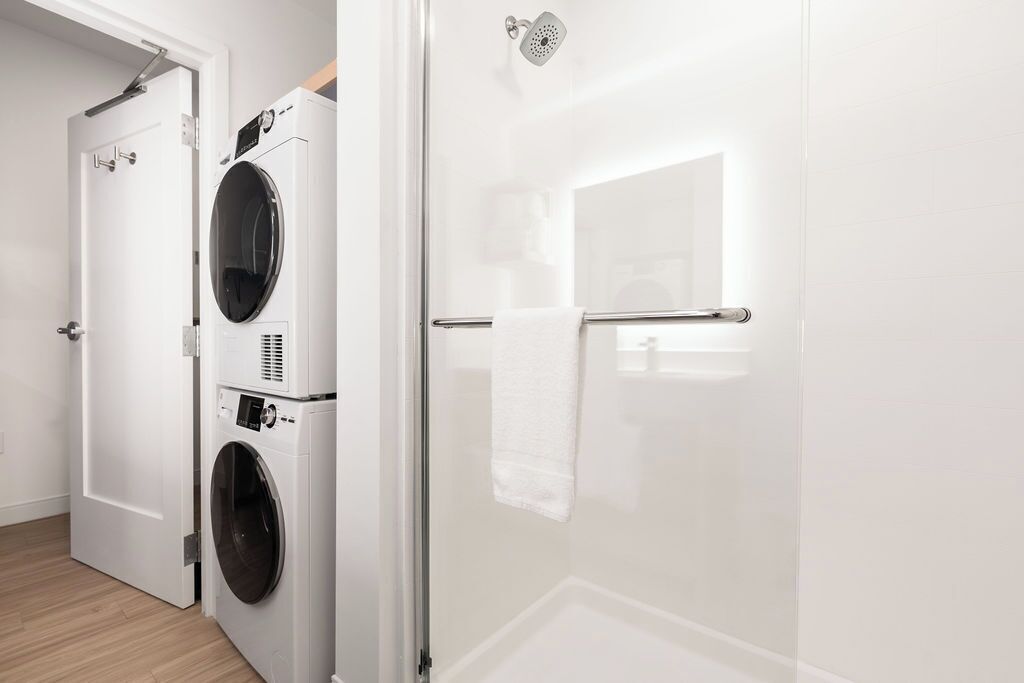 Une buanderie équipée d’un lave-linge et d’un sèche-linge, d’une cabine de douche en verre et d’une serviette blanche suspendue à un porte-serviettes.