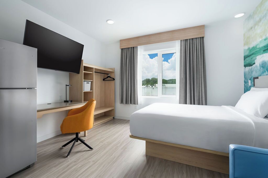 Una habitación de hotel con cama, escritorio, silla, televisión y ventana con cortinas.