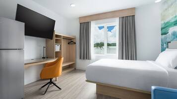 Una habitación de hotel con cama, escritorio, silla, televisión y ventana con cortinas.