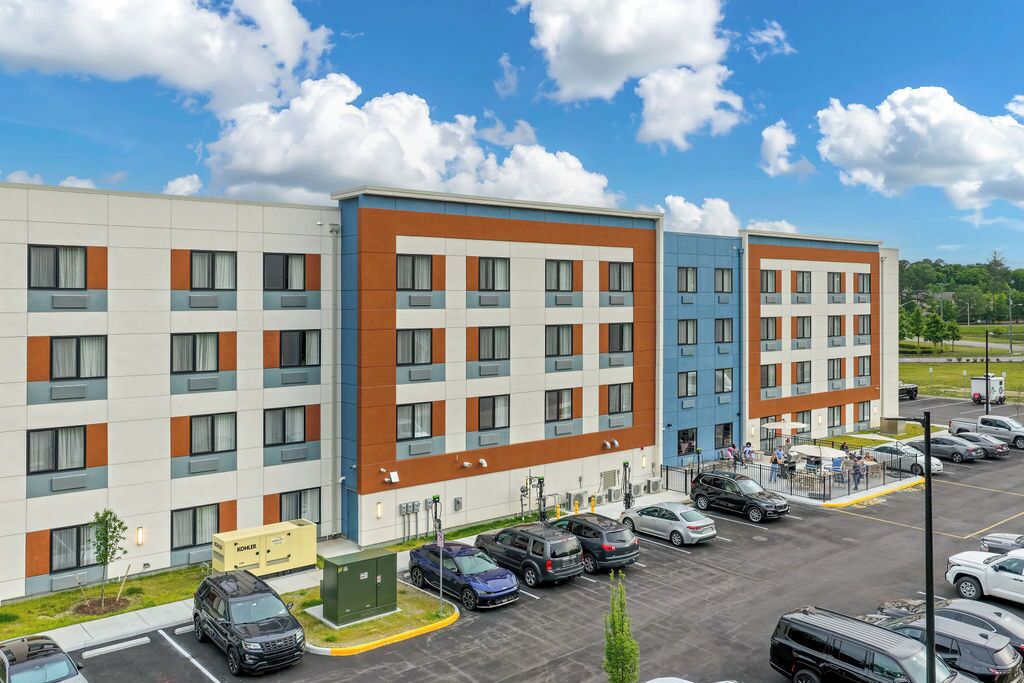 A multi-story hotel with a parking lot, a green area, and a clear sky.