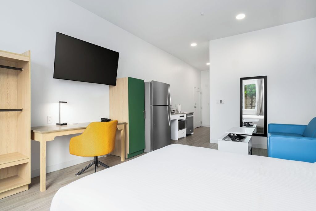 A modern hotel room with a bed, a desk, a chair, a TV, a kitchenette, and a window.