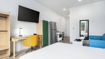 A modern hotel room with a bed, a desk, a chair, a TV, a kitchenette, and a window.