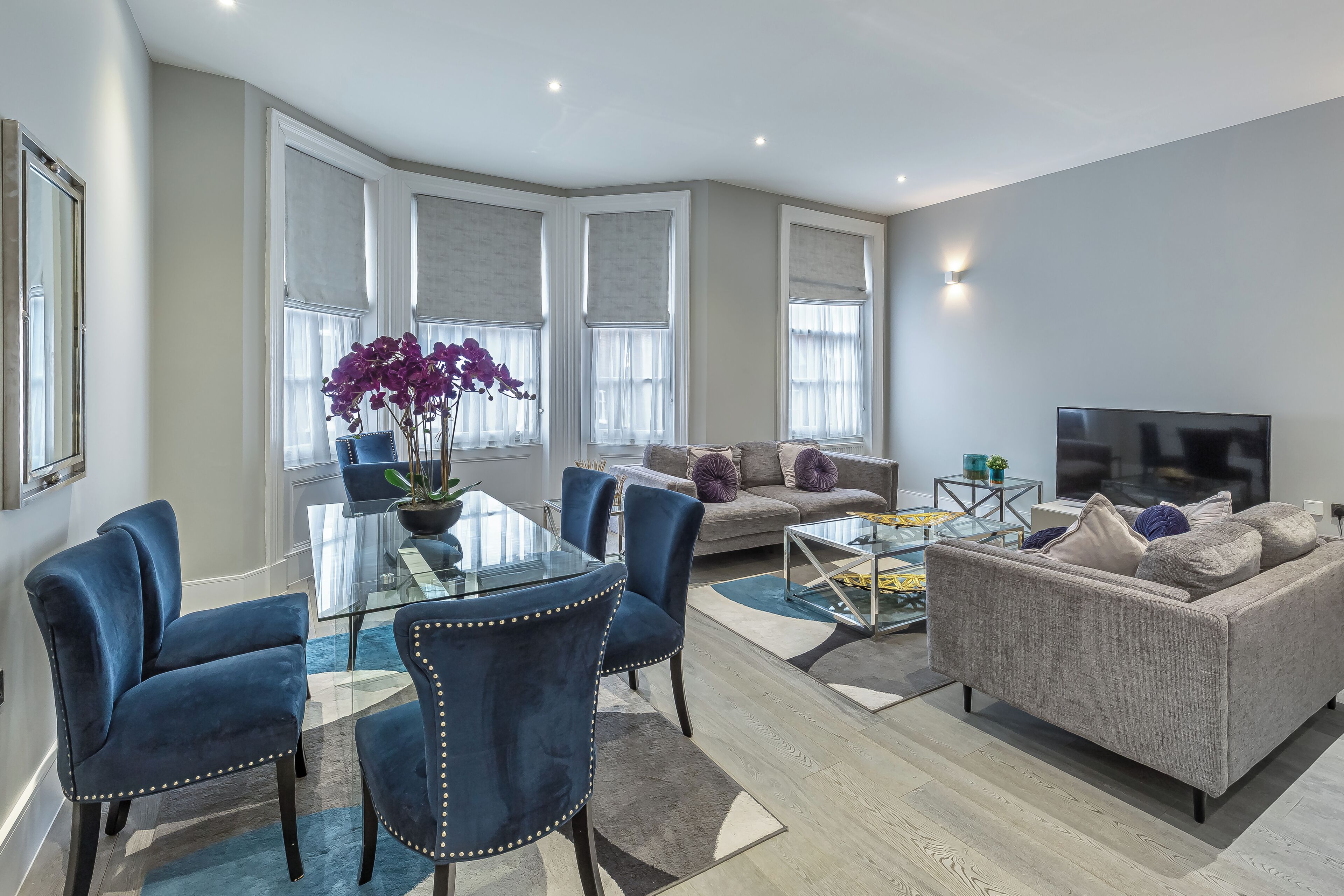 A modern living room with a glass dining table, blue upholstered chairs, a grey sofa, and a flat-screen TV.