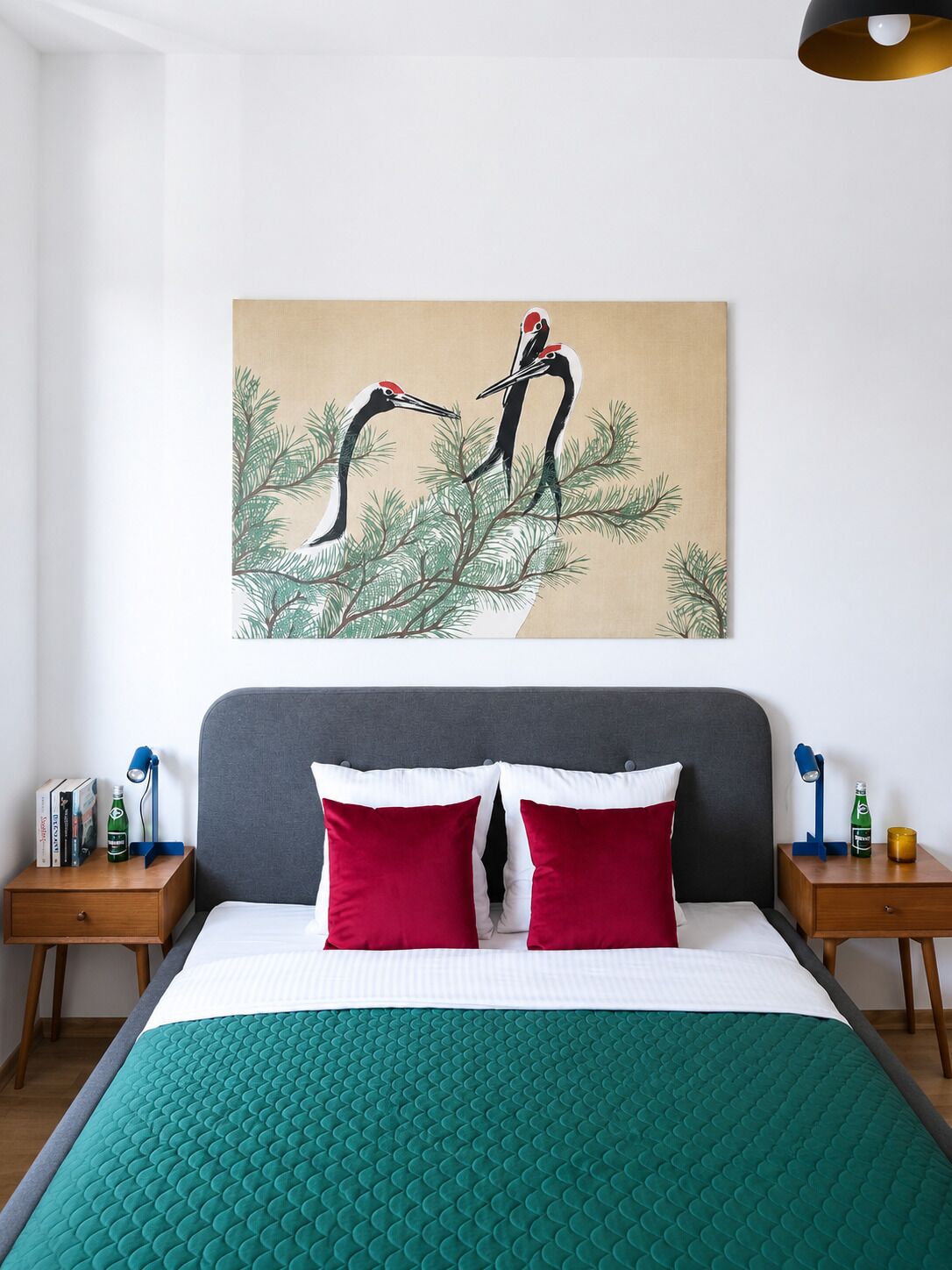 Une chambre à coucher avec un lit, deux oreillers rouges, une tête de lit grise, une œuvre d’art représentant des oiseaux accrochée au mur, et des tables de chevet avec des lampes et des bouteilles.