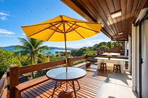 A wooden deck with a glass table and a yellow umbrella, overlooking a scenic view of the ocean and hills.