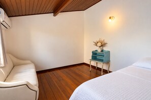 A hotel room with a wooden ceiling, a white bed, a white armchair, a small blue nightstand with a vase of flowers, and a wall-mounted light fixture.