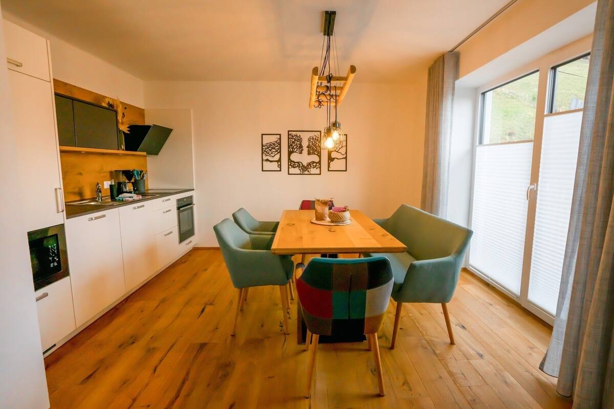 A modern kitchen with white cabinets, a wooden dining table, and four chairs. There is a pendant light above the table and a window with curtains to the right.